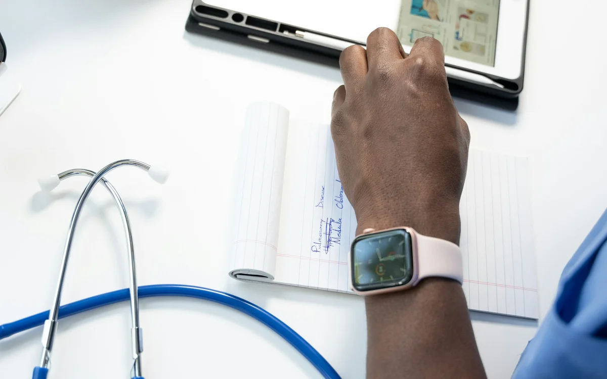 A doctor's hand writing notes beside a tablet and a stethoscope